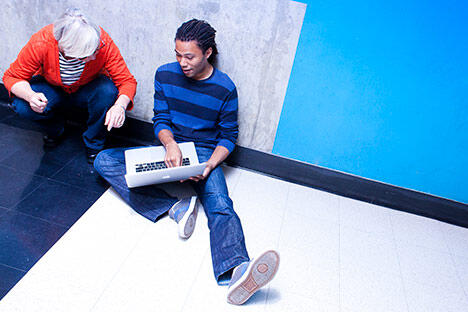 male student sitting on floor on the floor with an open laptop, talking with an older woman