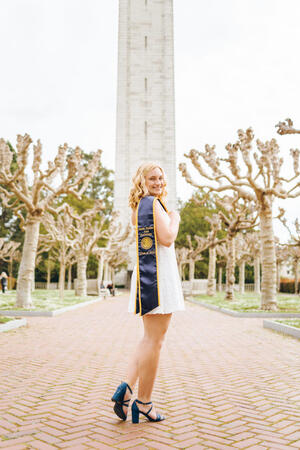 student wearing a white dress with a graduation sash standing in front of the campanile and smiling
