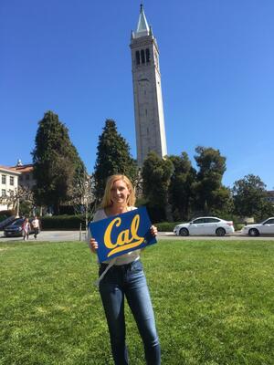 student standing on a grassy area holding a blue and yellow cal poster with the campanile in the background