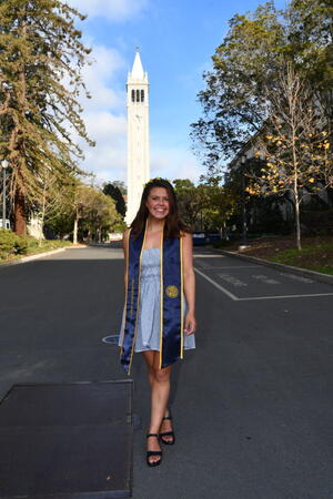 person standing in front of campanile smiling and wearing a graduation sash