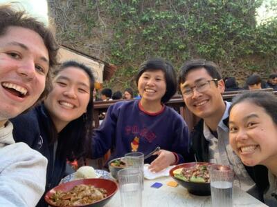 group of five students gathered around a table during the day eating food and smiling